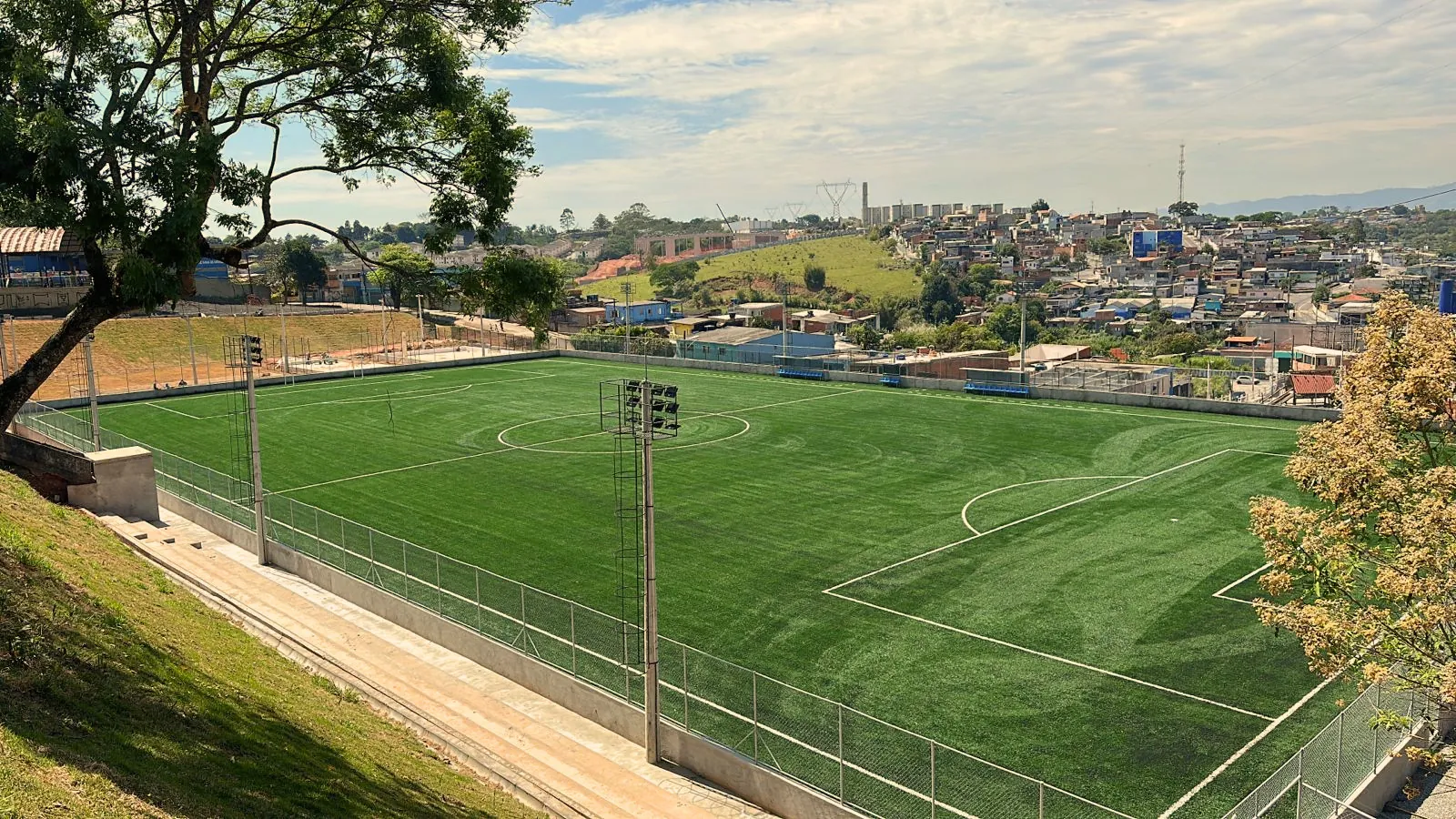 Vista elevada de campo esportivo em uso ativo, dez anos após a conclusão da obra, com bairro ao fundo