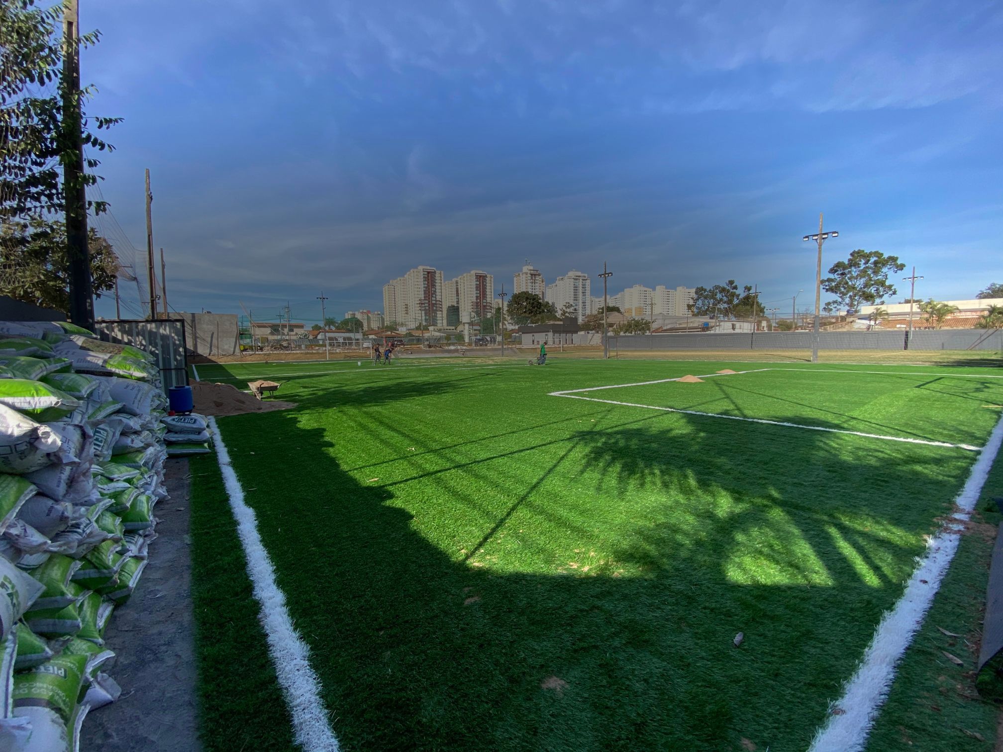 Campo esportivo em fase final de implantação, grama sintética recém-instalada, com skyline urbano ao fundo